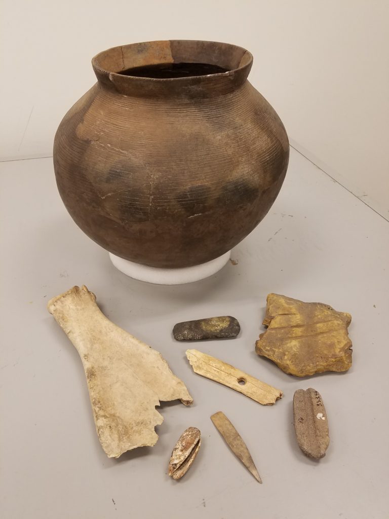The Sioux City Public Museum houses a collection of Kimball Village Site artifacts, including this grouping which features a restored clay vessel on loan from the State Archaeologist’s office. In the foreground, from left to right, are: a bison scapula hoe, an Olivella Shell pendant, a ground stone celt, a bone arrow shaft plane, a pointed bone awl, a grooved sandstone abrader for sharpening celts, axes and projectile points and a sandstone arrow shaft abrader. Photo courtesy of Sioux City Public Museum