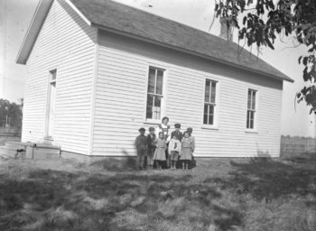 A group portrait of seven rural students with their teacher standing alongside their one-room schoolhouse in Muscatine County in 1910. The clapboard sided building has three tall windows, as well as wooden steps and a porch that lead to a single-wide doorway with a transom window. A field of corn can be seen behind the schoolyard. Photo by Oscar Grossheim. Photo courtesy of Musser Public Library